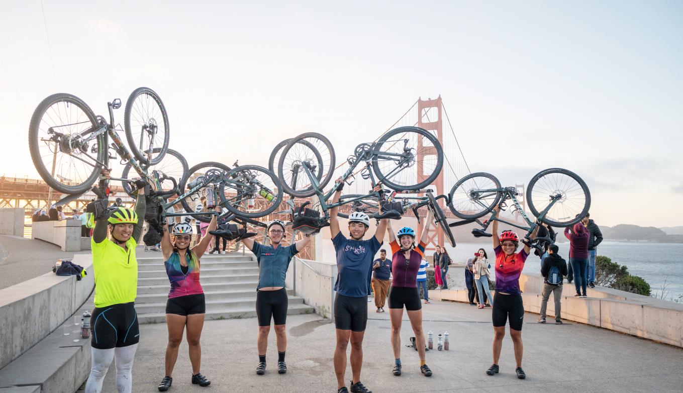 Six bicyclists in full riding gear stand in front of the Golden Gate Bridge holding their bicycles above their heads in triumph