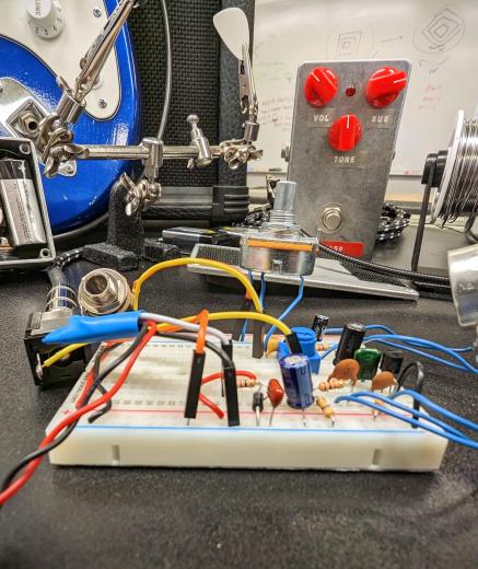 prototyping breadboard with components on bench if front of a blue guitar, opened guitar pedal with circuit exposed, and plain metal case pedal